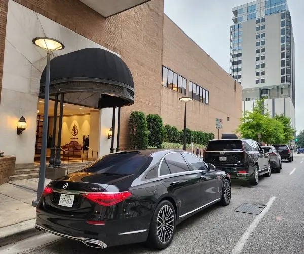 A sleek black Mercedes-Benz S-Class sedan parked under a black dome awning at a luxury hotel entrance. Other premium SUVs are lined up behind it on a city street, showcasing professional chauffeur pickup services.