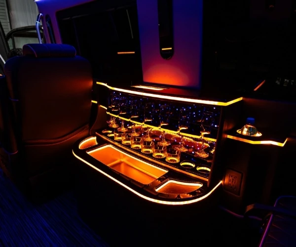Interior of a luxury limousine featuring a built-in bar with glowing orange neon accent lighting. The bar is stocked with crystal glassware and champagne flutes.