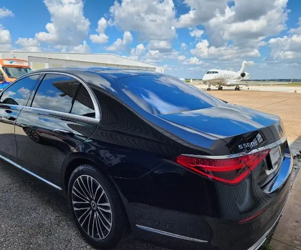 Rear view of a black Mercedes-Benz S500 parked on an airport tarmac next to a private white jet. The car has a polished finish reflecting the blue sky and clouds, emphasizing premium airport transfer services.